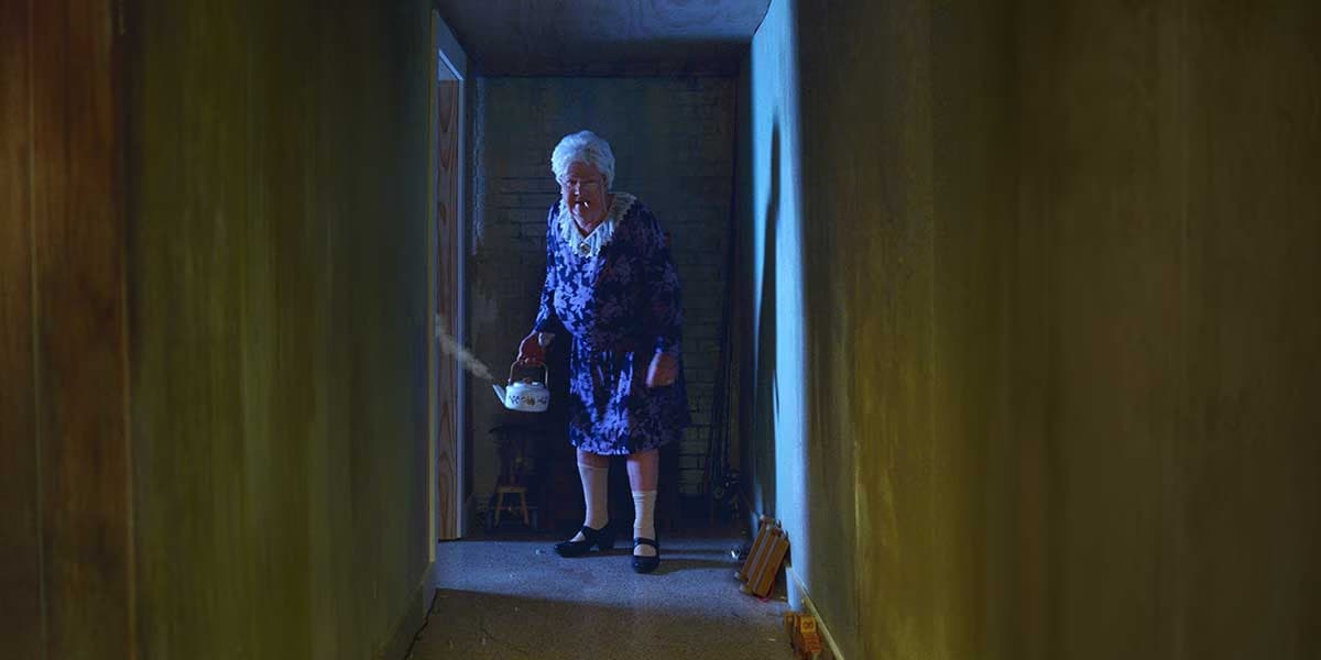 An elderly woman in a patterned dress stands holding a white kettle in a dim narrow hallway.