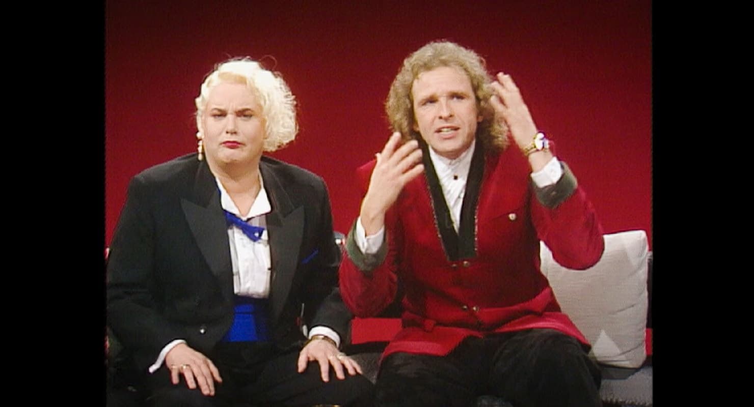 Two presenters in formal attire sit against a solid red background during a television broadcast.