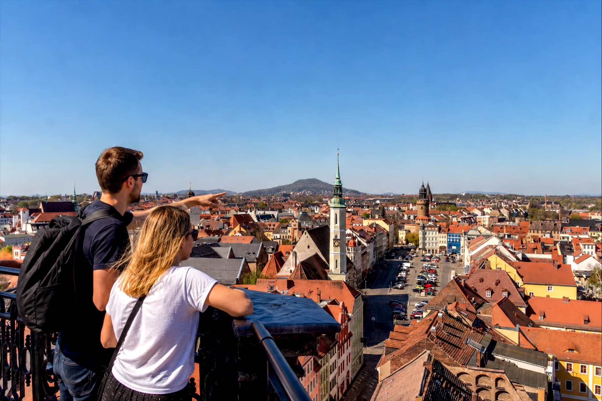 A young couple standing on a viewing platform looks over a historic European town with red roofs.