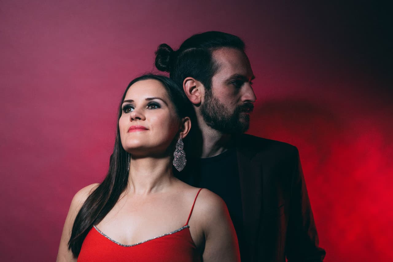 A woman in a red top and a man in a black shirt pose against a reddish background.