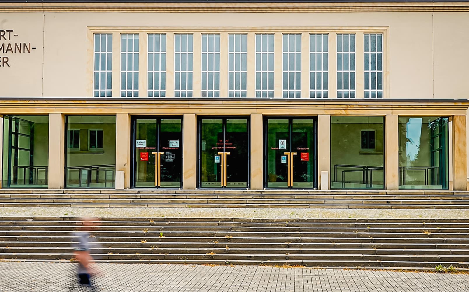 Front facade of a public building with stairs and a blurred person walking by.