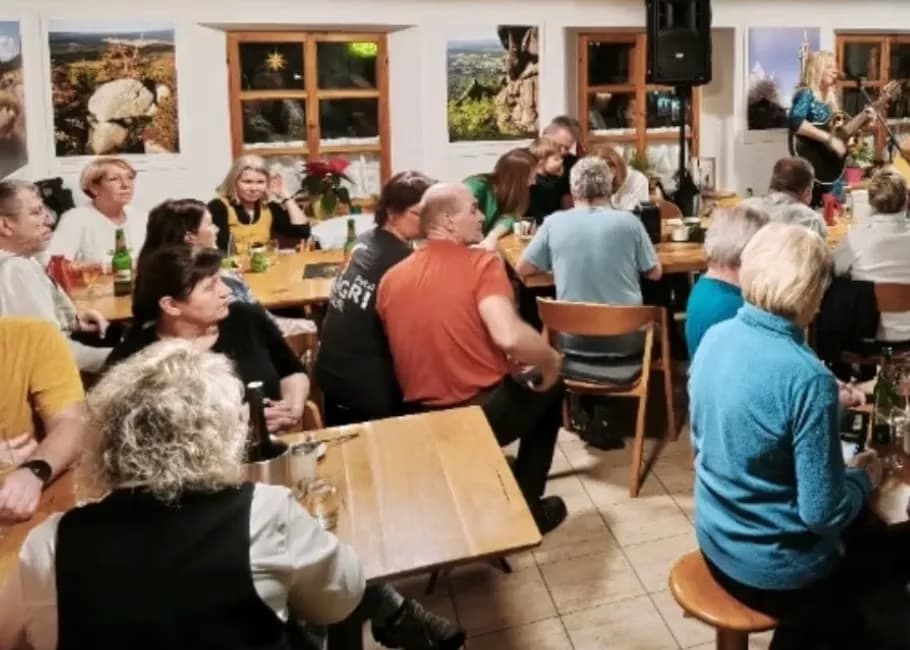 Leute sitzen in einem rustikalen Raum an Tischen, während eine Frau in der Ecke Musik macht.