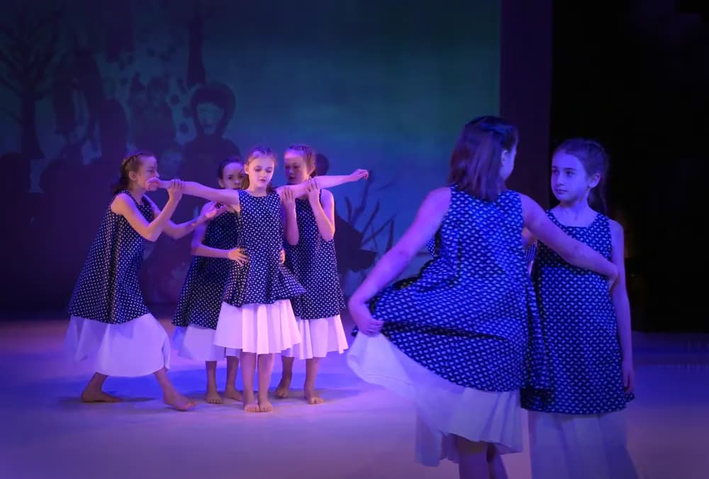 Junge Mädchen in blau gepunkteten Kleidern tanzen auf einer blau beleuchteten Theaterbühne.