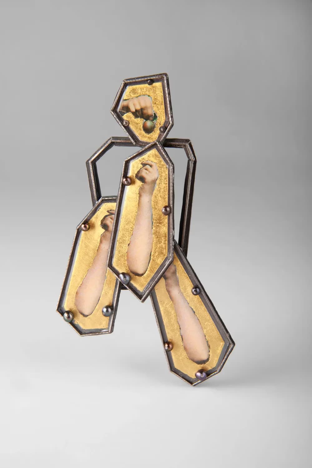 Abstract brooch made of geometric metal frames containing gold leaf and miniature images of human arms.