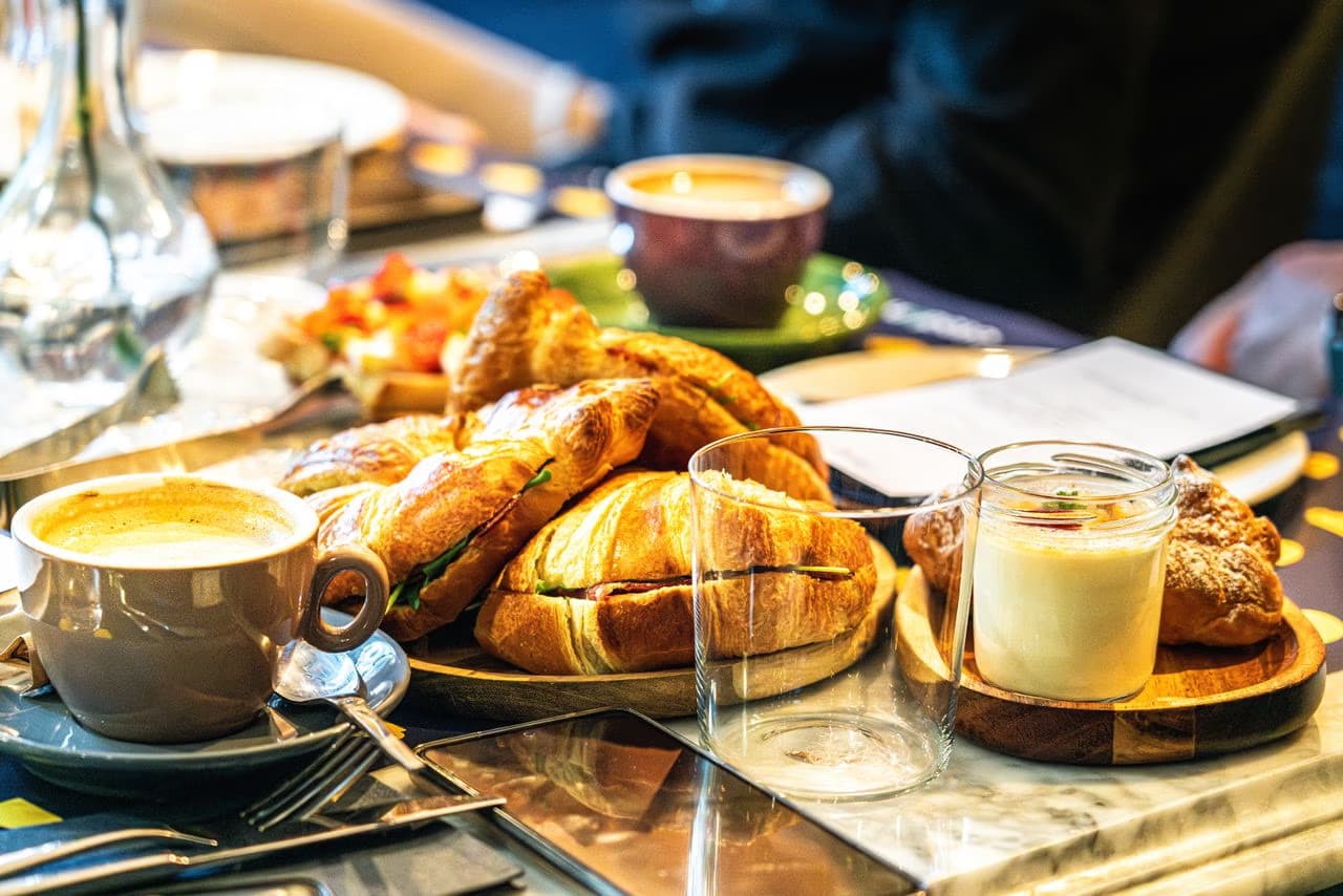 A breakfast table spread featuring filled croissants, fresh coffee, and glassware at a cafe.