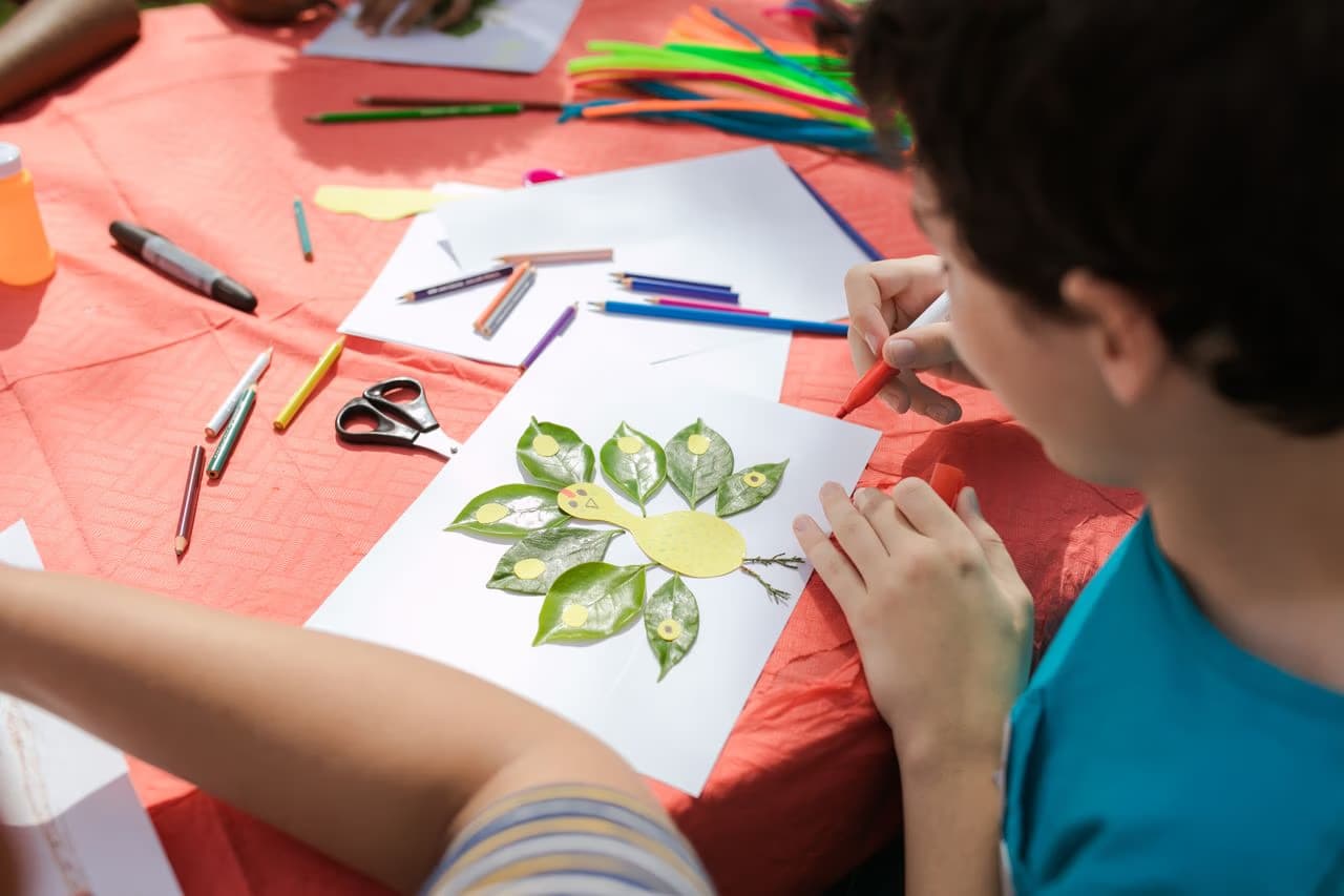 A student works on a craft project using leaves and markers at a red table.