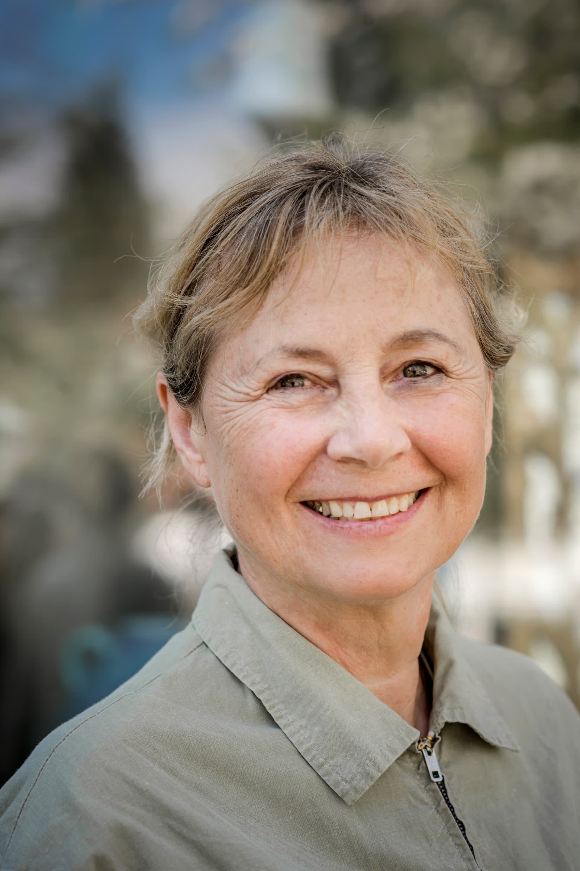 A close-up portrait of a smiling middle-aged woman with light brown hair outdoors.