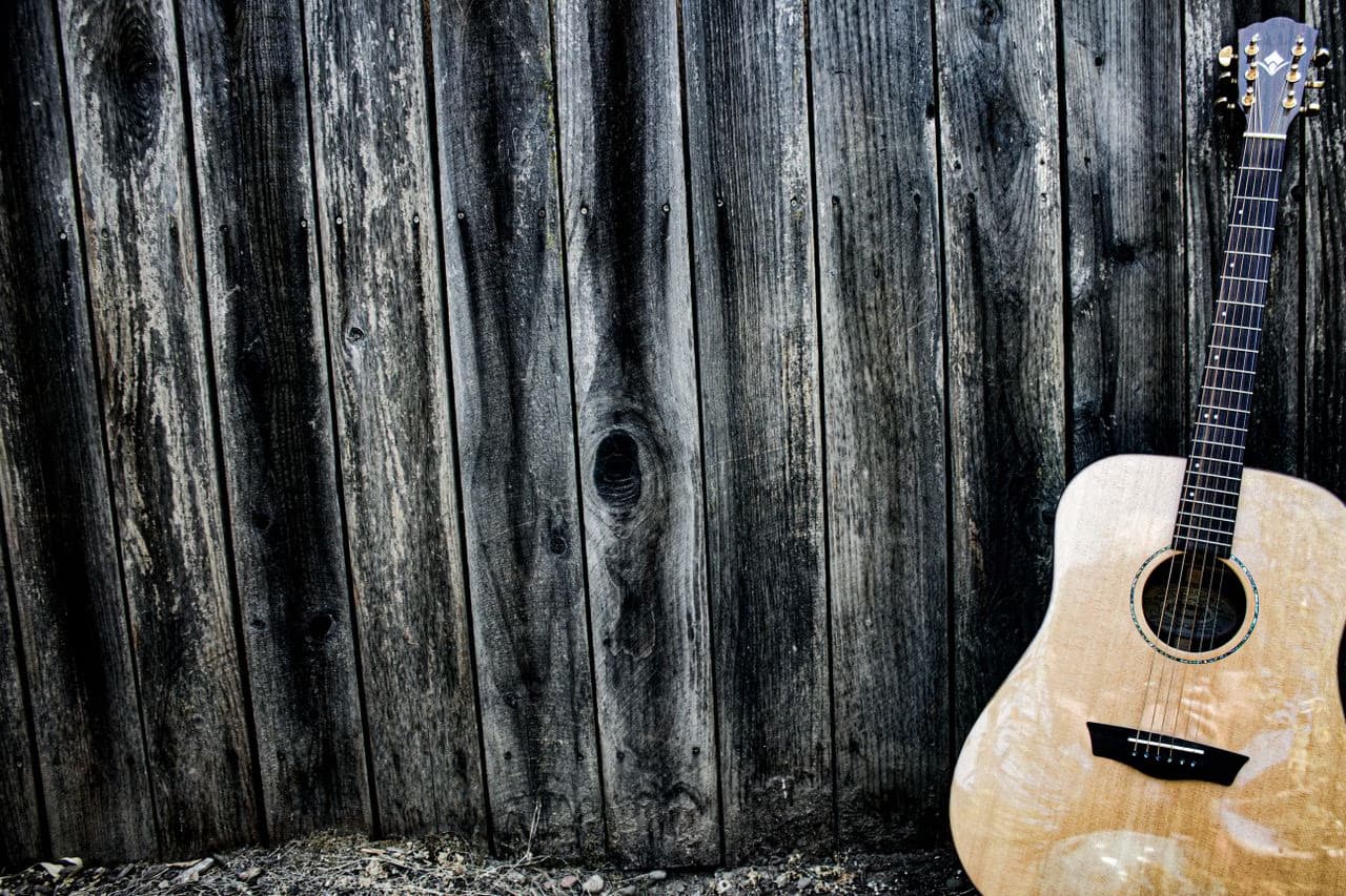 A light-colored acoustic guitar leans against a weathered dark wooden fence.