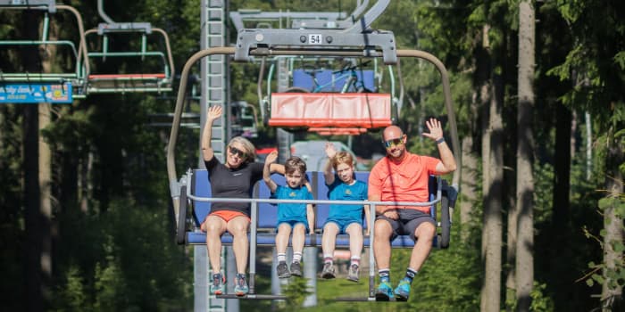 A family of four waving while riding an open-air chairlift through a green forest.