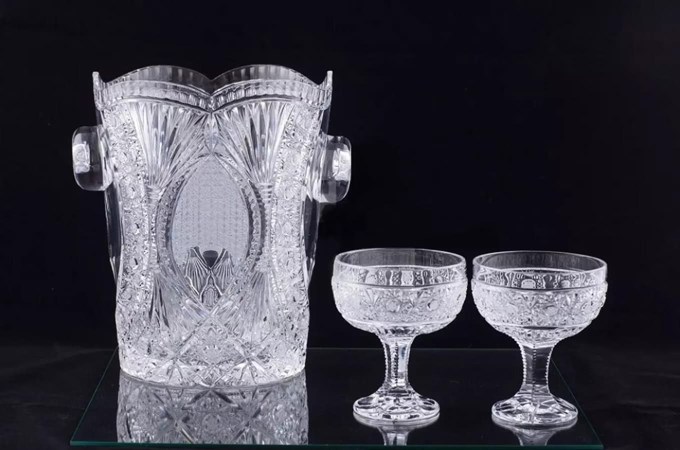 A cut glass ice bucket and two matching stemmed glasses on a reflective surface.