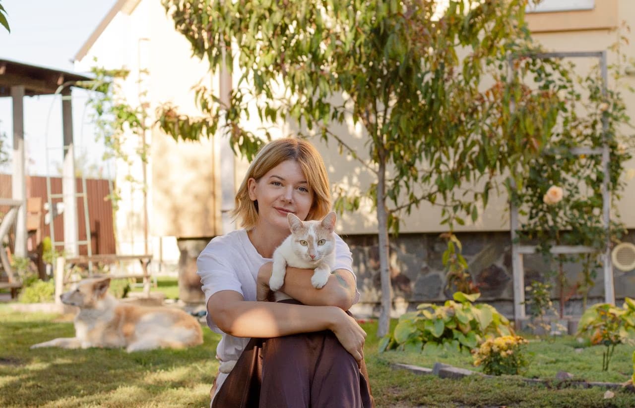 A young blonde woman sitting in a sunny garden holding a cat with a dog resting nearby.