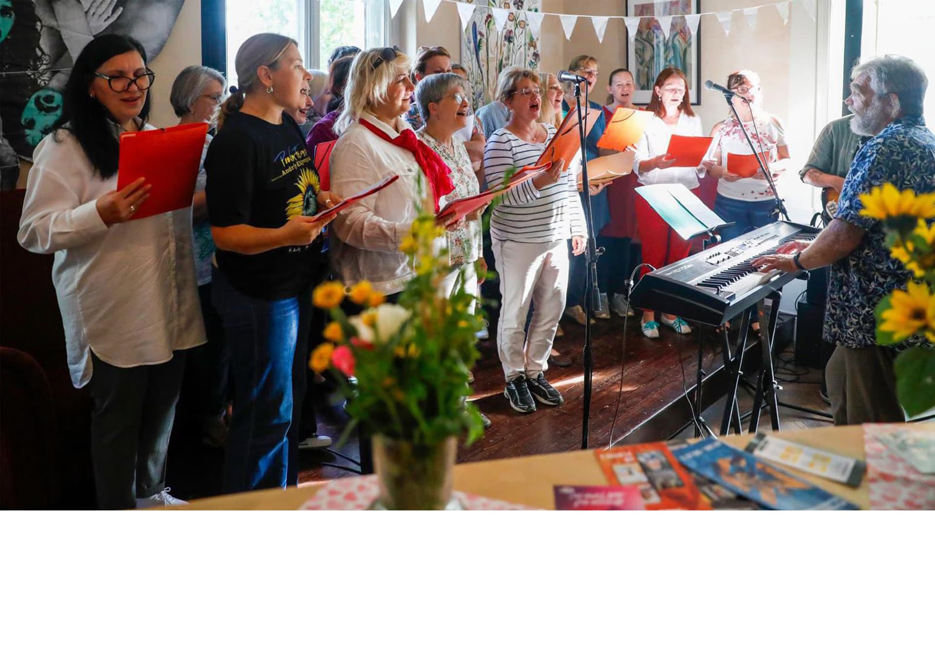 A diverse group of singers stands in a room, holding red folders while performing with a keyboardist.