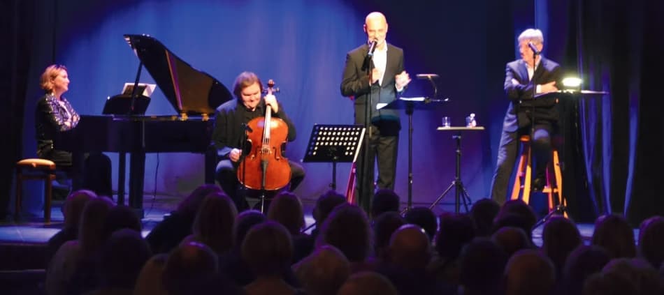 Musiker und ein Redner treten vor einem Publikum in einem schwach beleuchteten Saal auf.