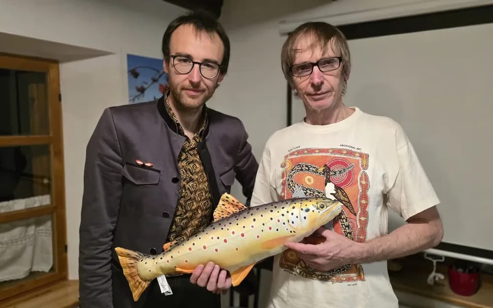 Two men standing indoors holding a large stuffed brown trout plush toy.