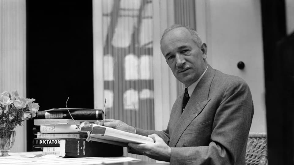 A middle-aged man in a suit sits at a desk beside a stack of books labeled Dictator.