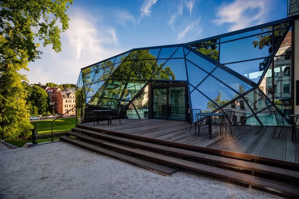 Modern angular glass building with a wooden terrace in a park setting.