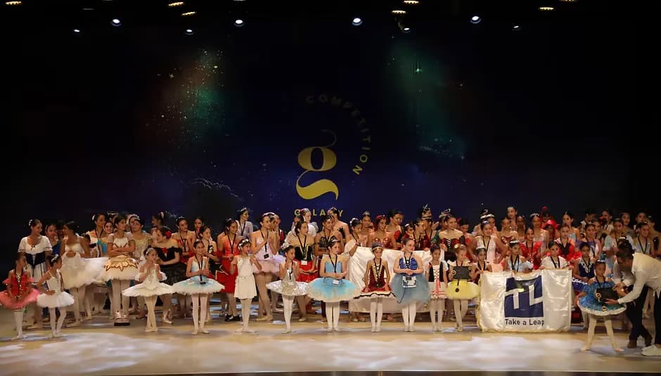A large group of ballet dancers in various costumes poses on a brightly lit theater stage.