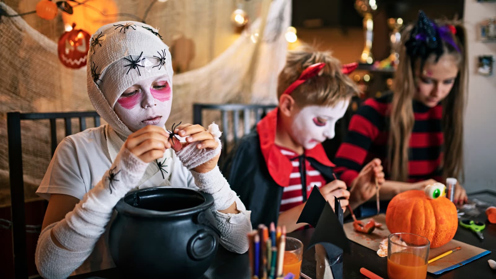 Děti v halloweenských kostýmech pracují u stolu na strašidelných řemeslných výrobcích.