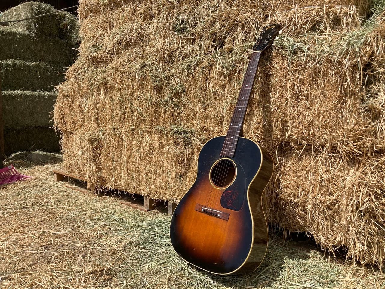 A sunburst acoustic guitar leans against a wall of stacked hay bales inside a rustic barn.