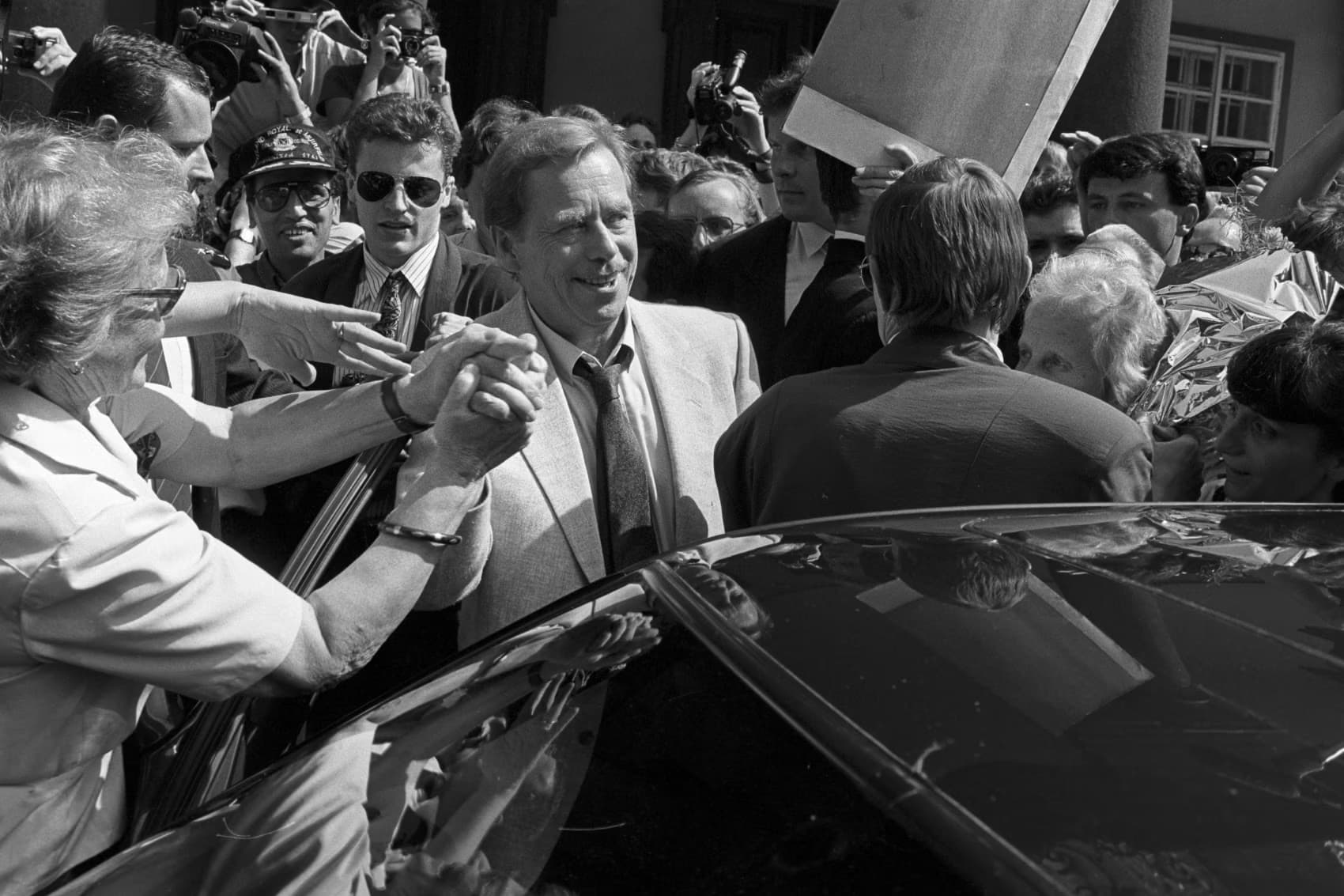 Vaclav Havel smiles while surrounded by a crowd of people in a busy street setting.