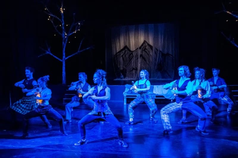 Performers in blue stage lights dance on a dark theater stage with mountain silhouettes.