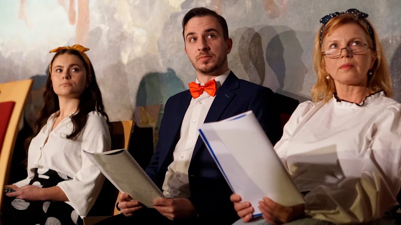 Three people holding scripts sit in a row during a formal rehearsal or reading.