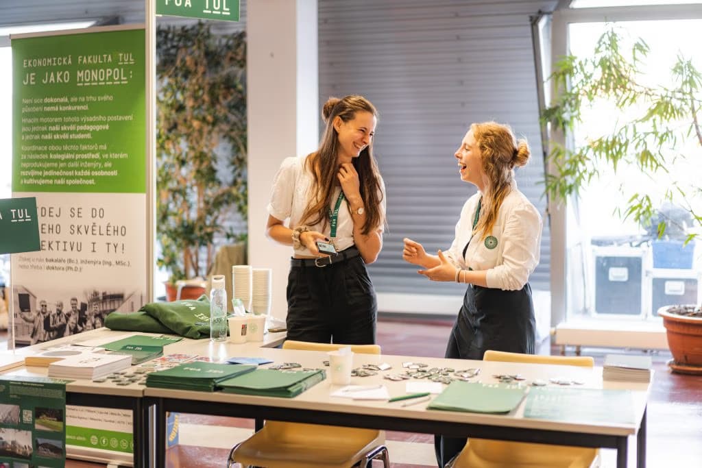 Two young women in white shirts laugh together while standing at a promotional booth.