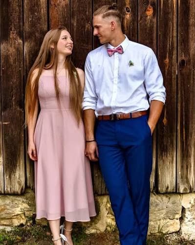 A young couple in formal attire stands together against a rustic wooden wall.