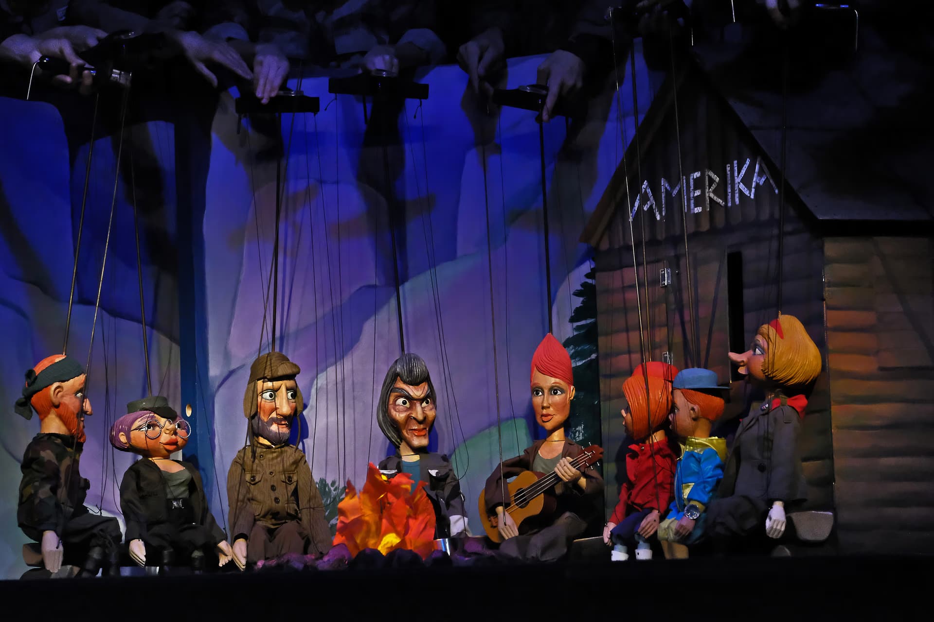 Several marionettes sit around a campfire on a stage in front of a wooden house labeled AMERIKA.