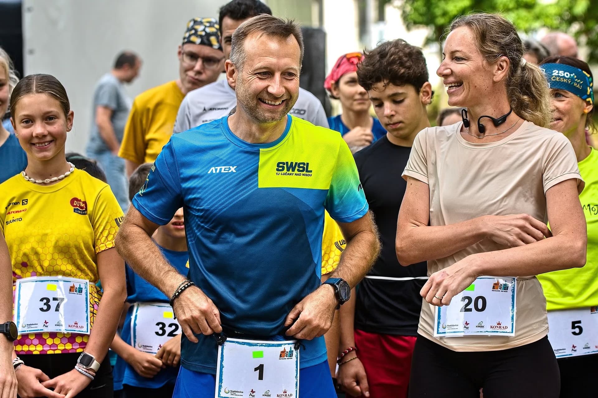 Smiling runners of various ages stand together wearing race bibs at an outdoor event.