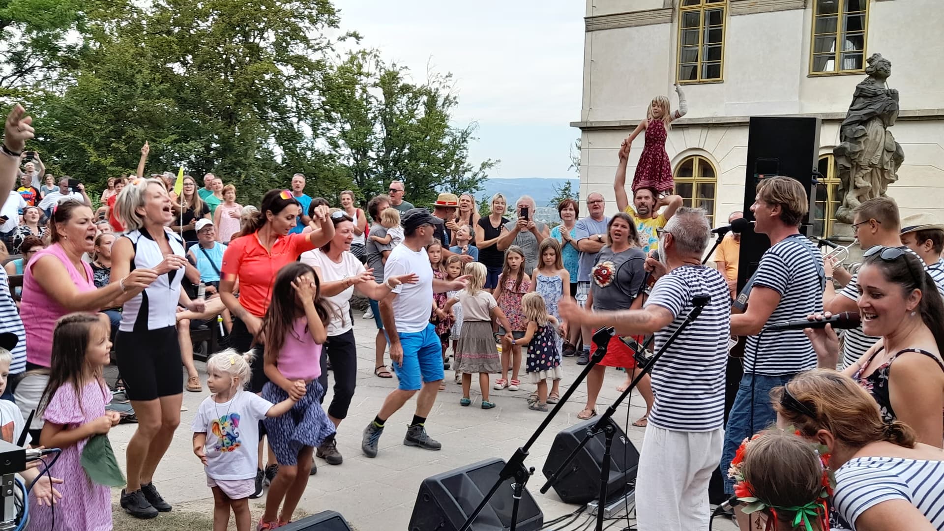 A crowd of adults and children dancing outdoors in front of a building during a musical event.