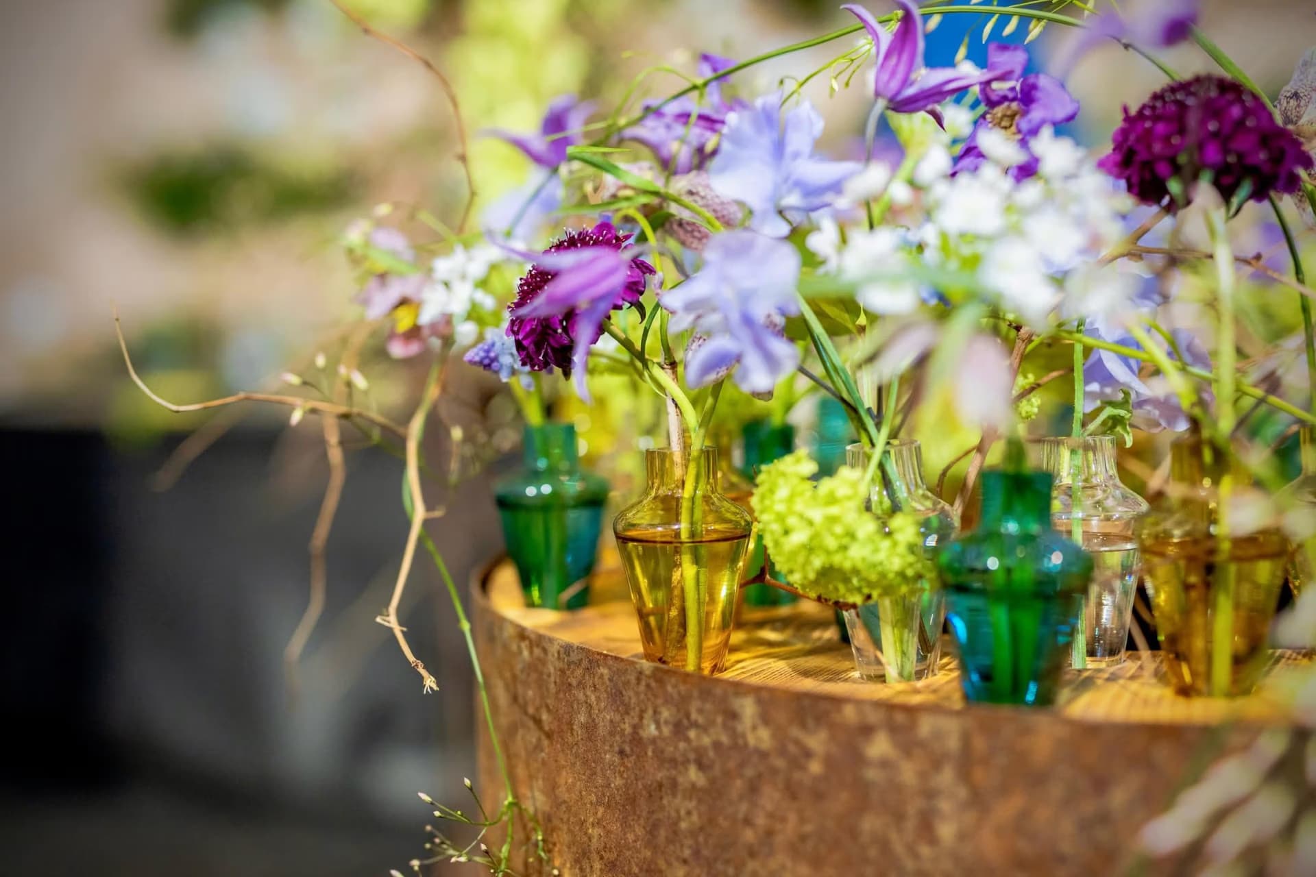 Several small colorful glass vases containing delicate spring flowers arranged on a weathered circular base.