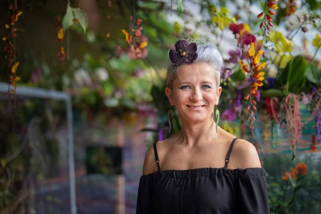 Smiling woman in black top with silver hair and flower accessory stands among hanging orchids.