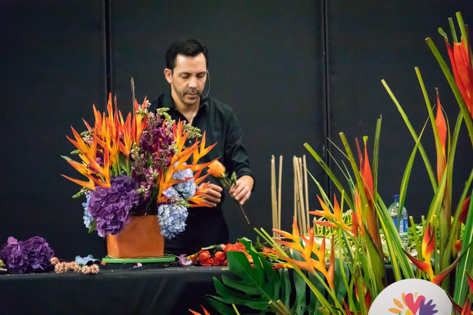 A florist arranges colorful tropical flowers in a bouquet while standing at a black table.