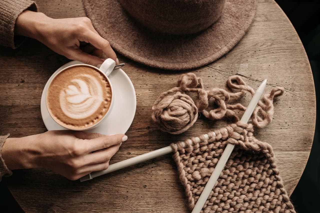 Hands hold a cup of latte beside knitting needles and wool on a wooden table.