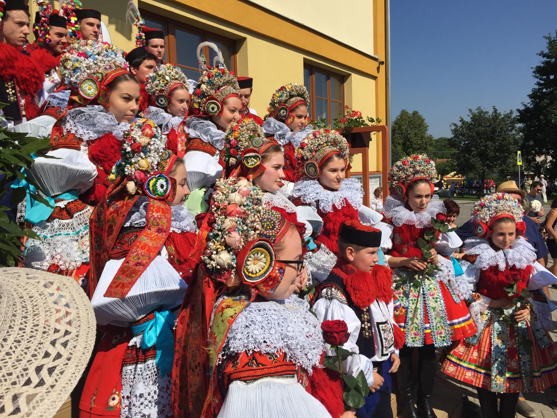 A group of people standing outdoors wearing traditional colorful folk costumes with elaborate floral headpieces.