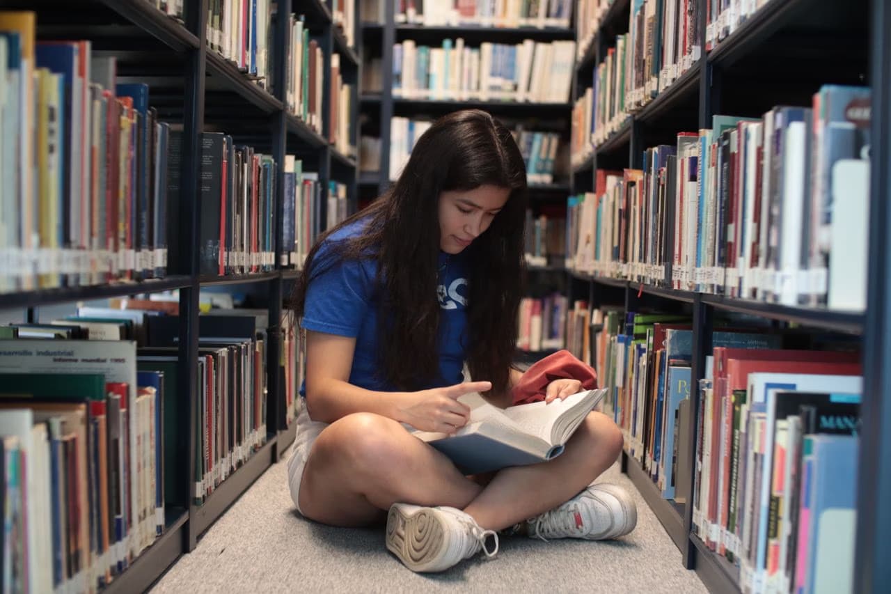 A student in a blue shirt sits cross-legged on the floor reading in a library aisle.