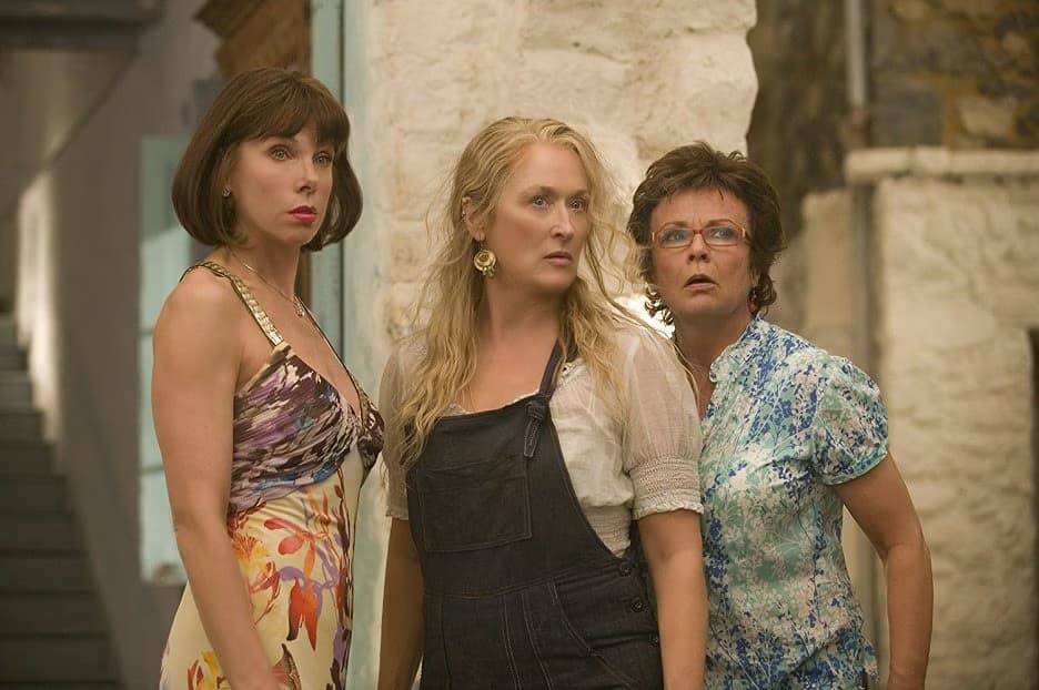 Three women stand outdoors in front of a white stone wall looking surprised.