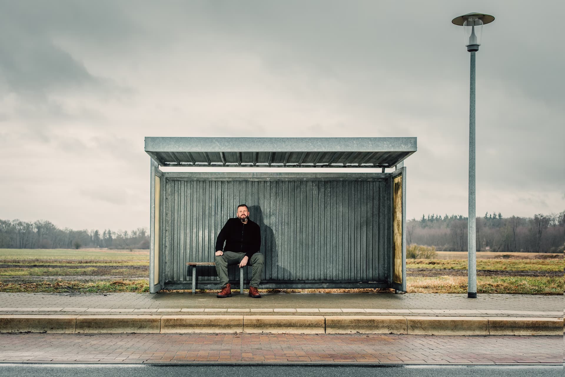 A man sits alone inside a metal bus shelter on a rural road under an overcast sky.