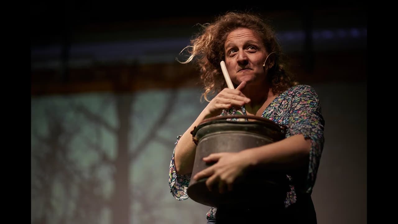 A curly-haired woman on stage holds a metal bucket and stick in front of a tree backdrop.