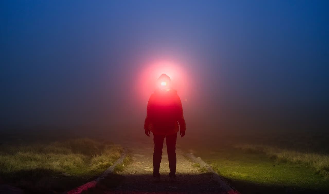 A hiker stands on a dark, misty trail with a bright red headlamp beam illuminating the fog.