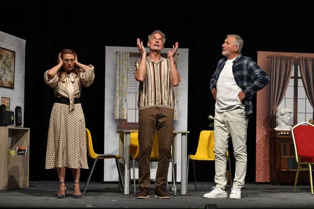 Three actors perform on a theater stage set with chairs and a table against a black background.