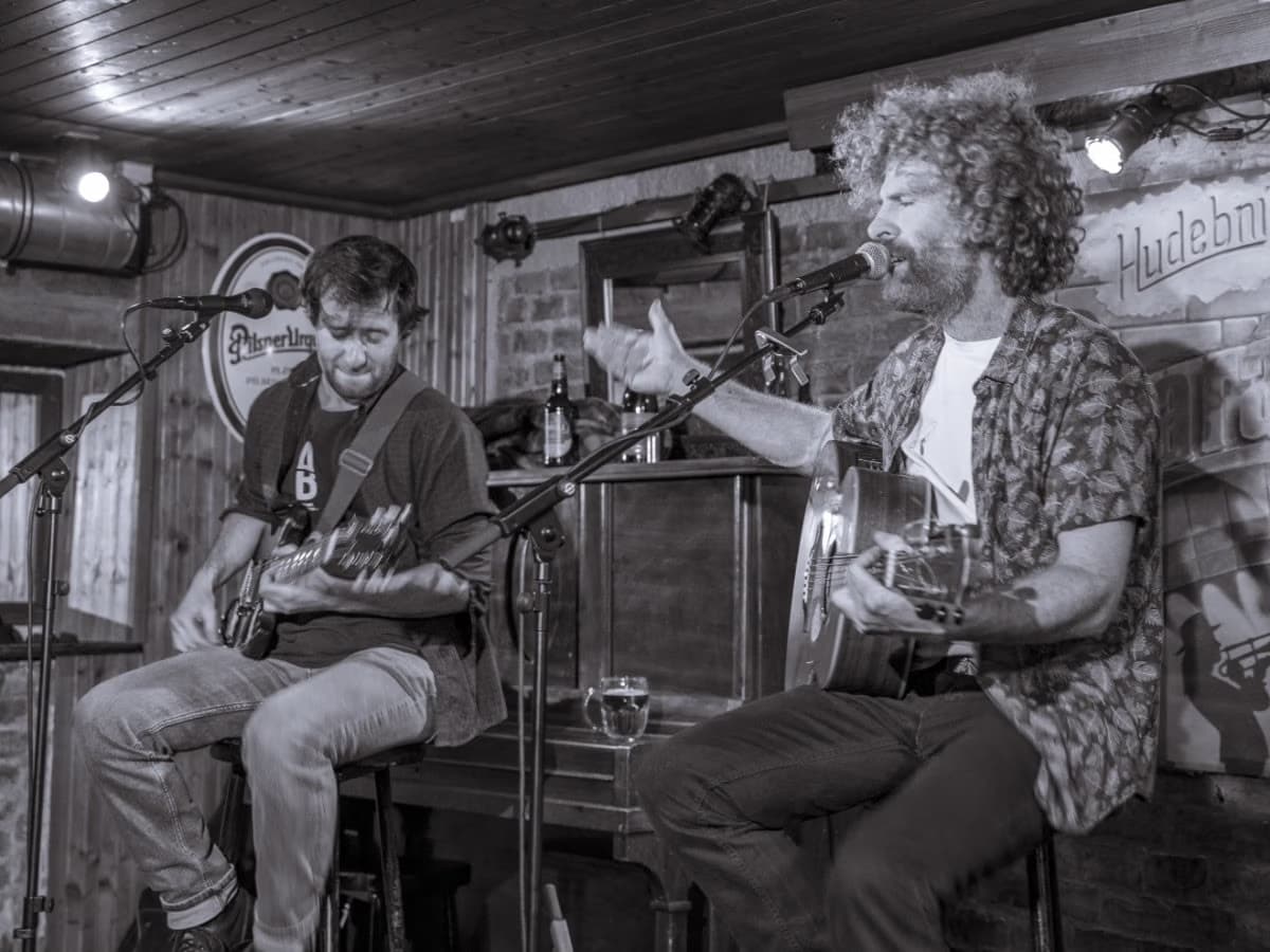 Zwei Männer spielen Akustikgitarren und singen in einem beleuchteten Pub in Mikrofone.