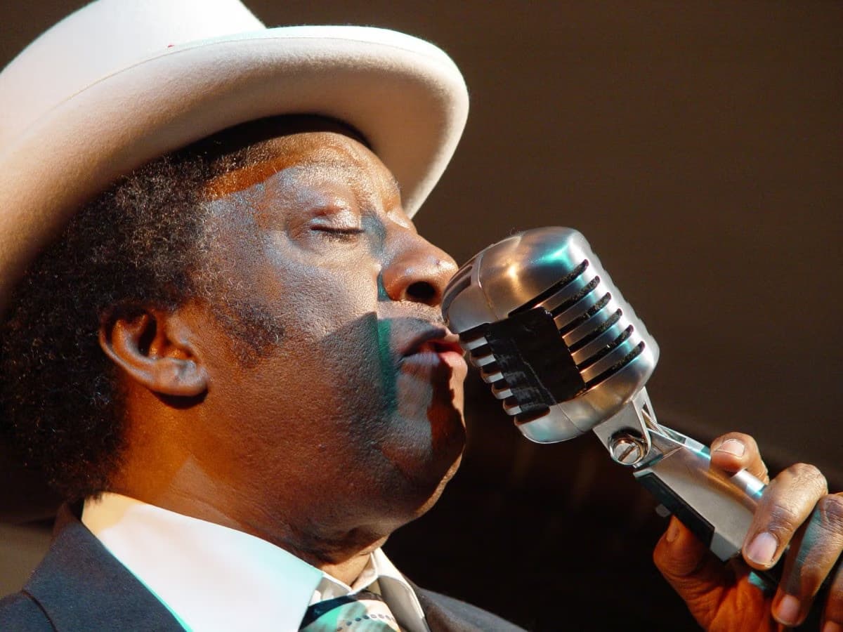 Ein afroamerikanischer Mann mit Hut singt leidenschaftlich in ein altes silbernes Mikrofon.