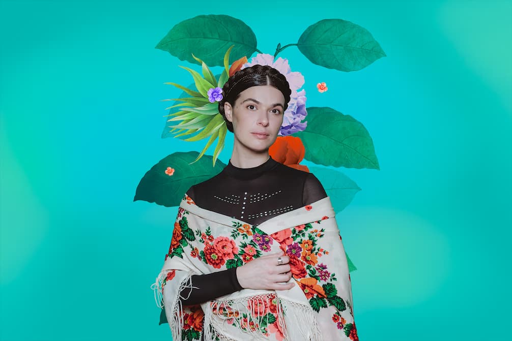 Eine Frau mit Blumenkranz und buntem Tuch posiert vor einem leuchtend türkisfarbenen Hintergrund.