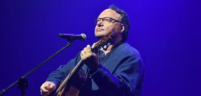 A bespectacled man in a blue shirt plays acoustic guitar and sings on stage under blue light.