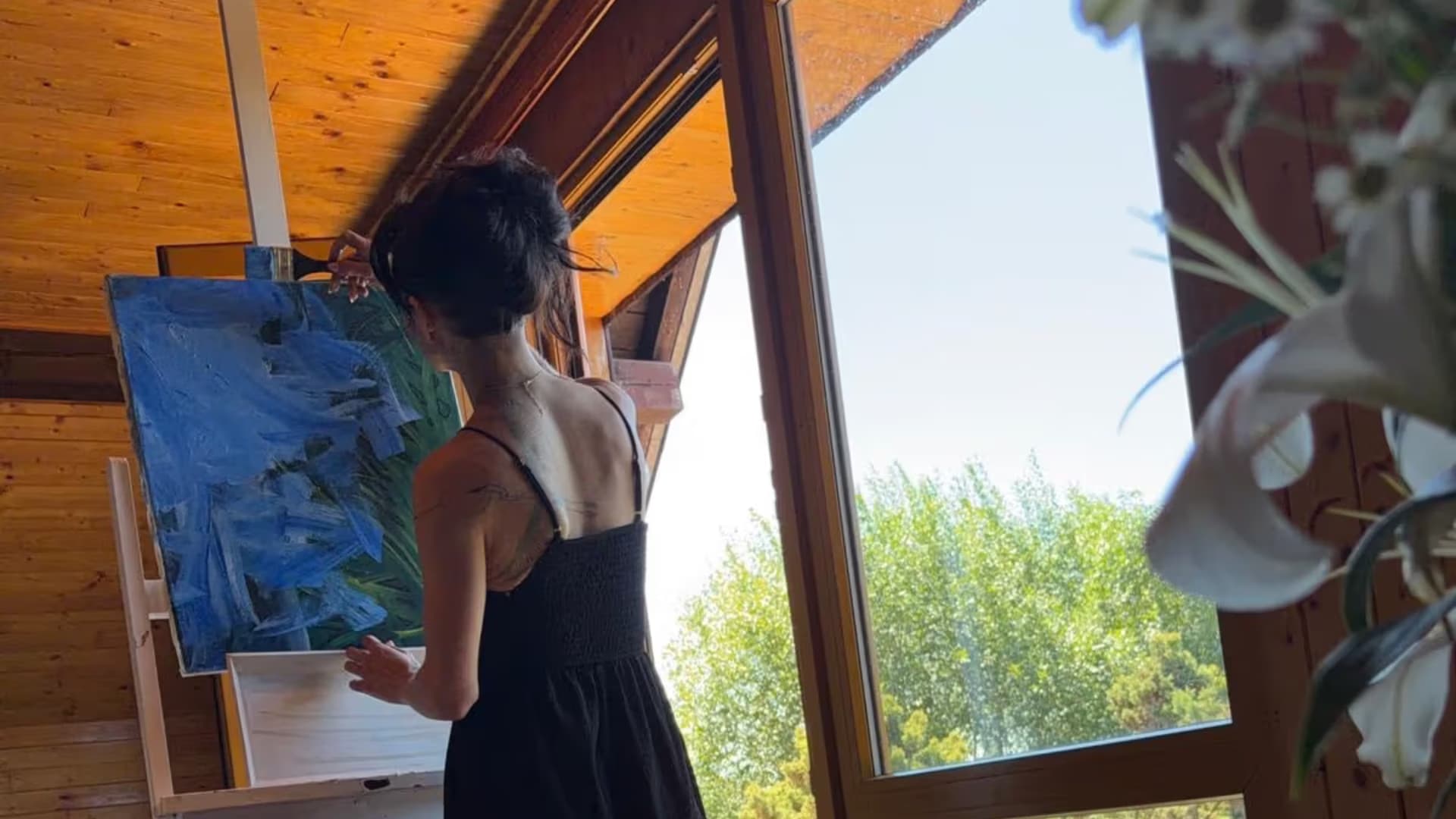 A woman in a black dress paints on a canvas inside a sunlit wooden room.