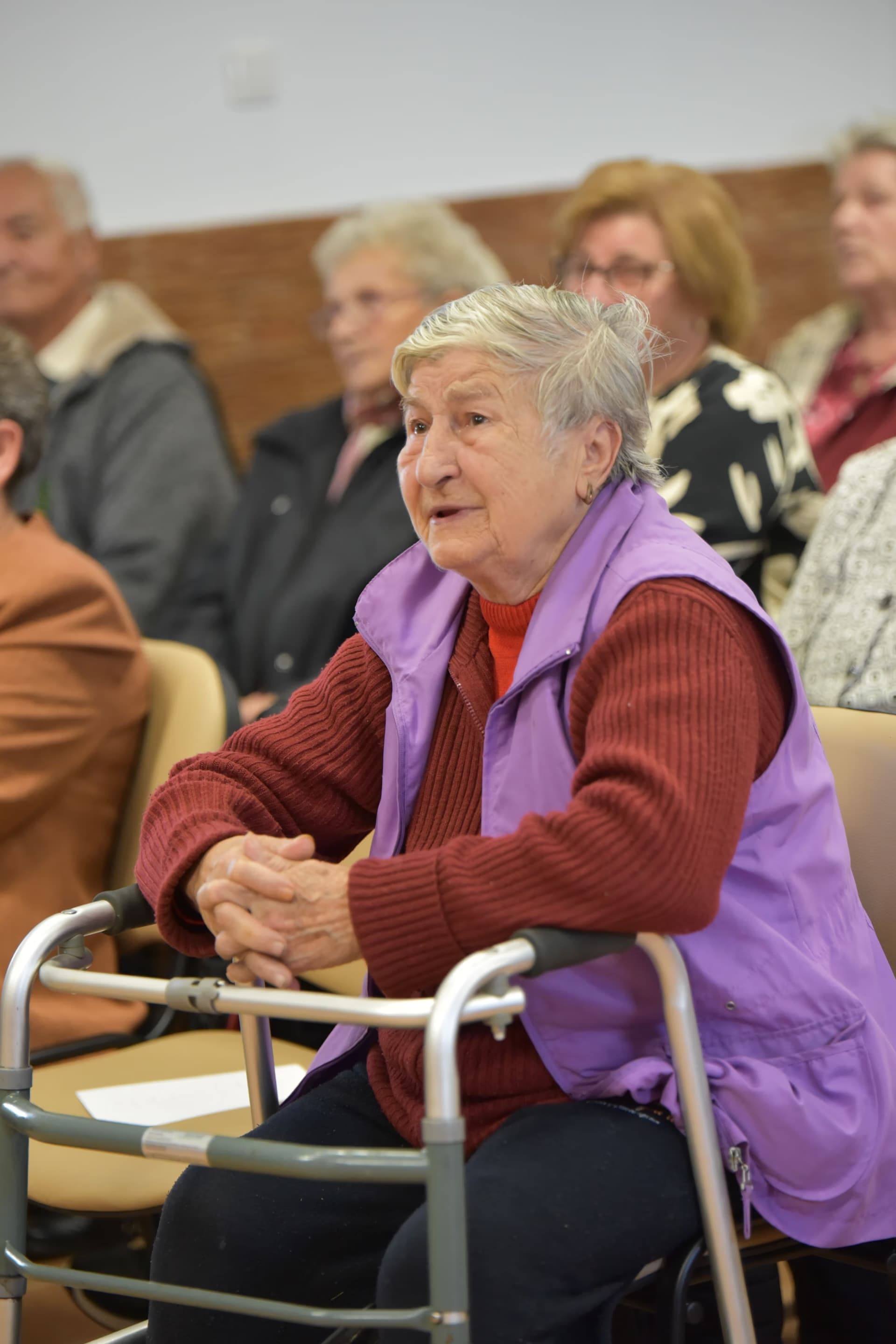 Elderly woman with a walker sitting in an audience among other people at a public event.