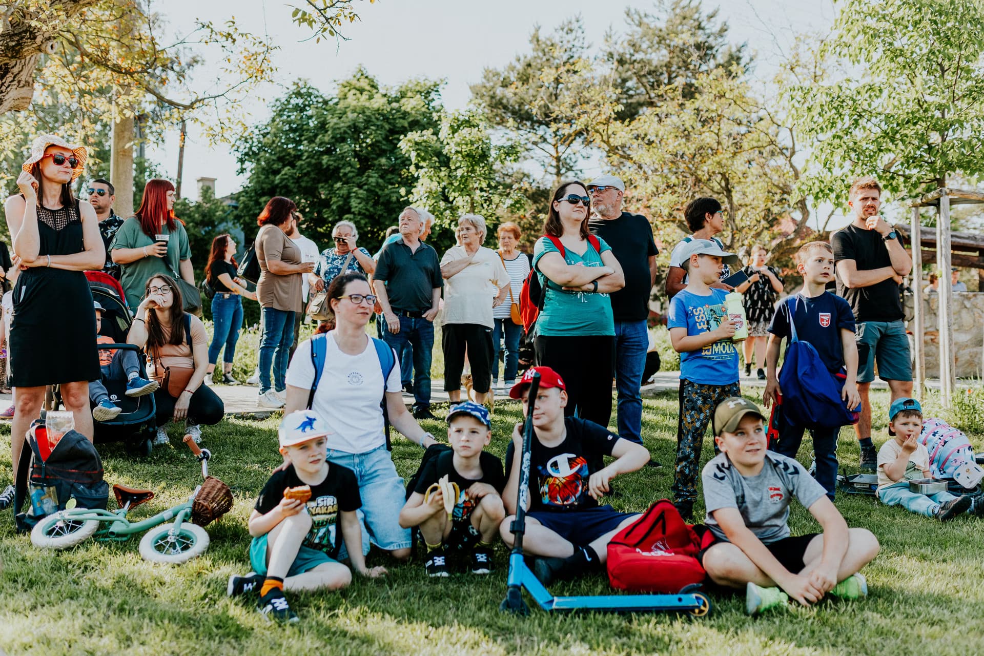 A diverse crowd of adults and children sit on grass at an outdoor event on a sunny day.