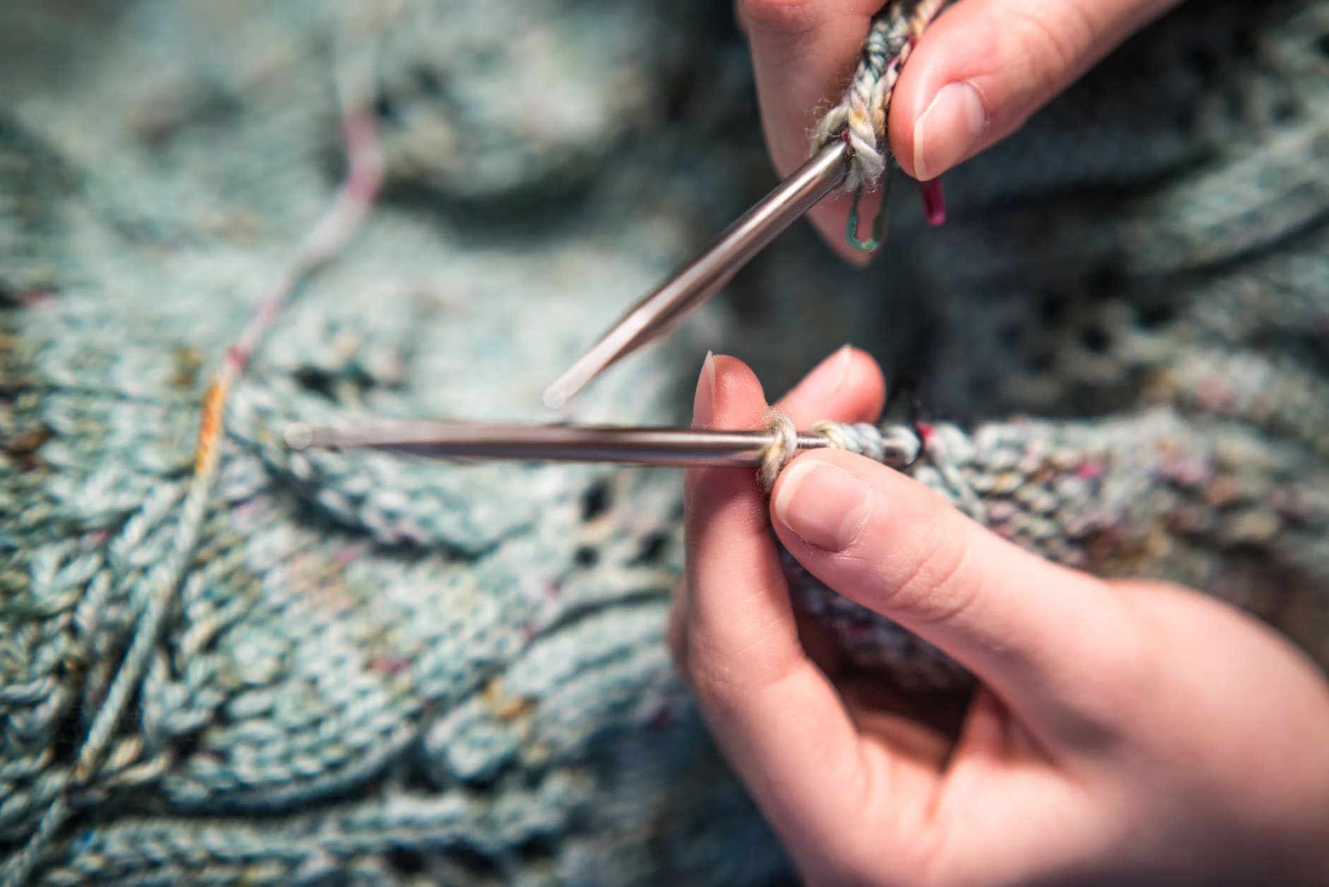 Close-up of hands knitting with metal needles and textured blue-grey yarn.